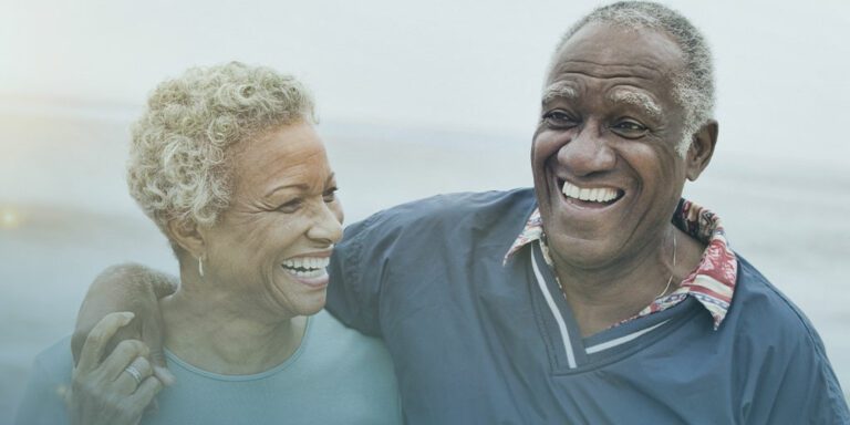a smiling older man with dark skin has his arm around a smiling older dark-skin woman as they celebrate healthy aging month